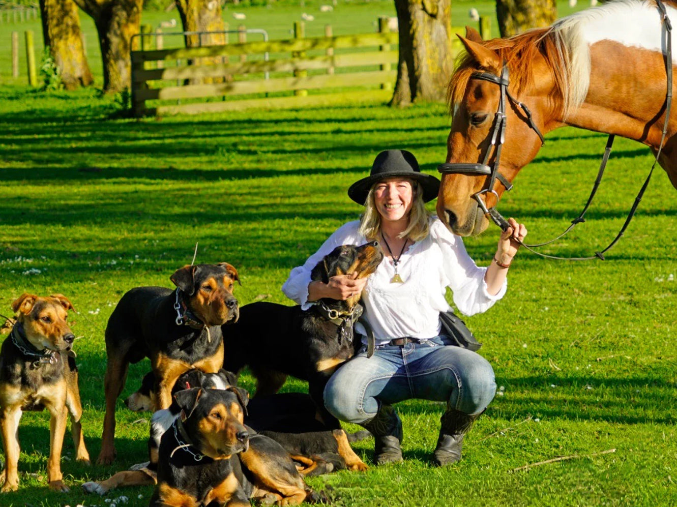 Woman posing with several dogs and a horse in a sunny green field.