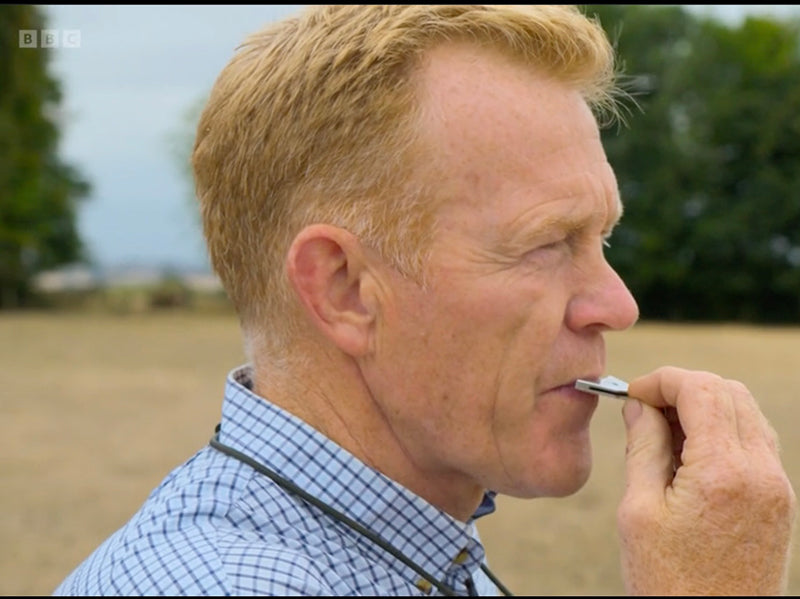 Man blowing a metal dog whistle outdoors in a field.