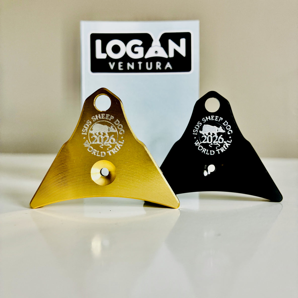 Two Logan Ventura sheepdog whistles, one gold and one black, engraved with the ISDS World Trial logo, displayed on a reflective white surface with a white Logan Ventura card in the background.