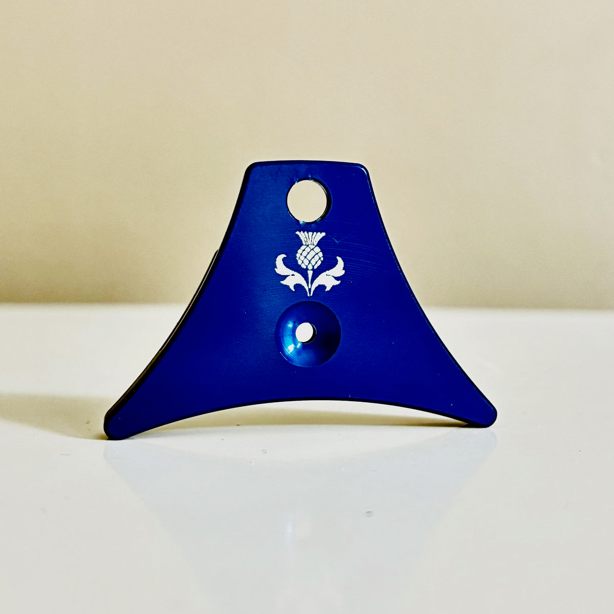 A blue anodized Logan A1 Blue sheep dog whistle awith a white engraved thistle logo, resting on a white surface against a beige background.